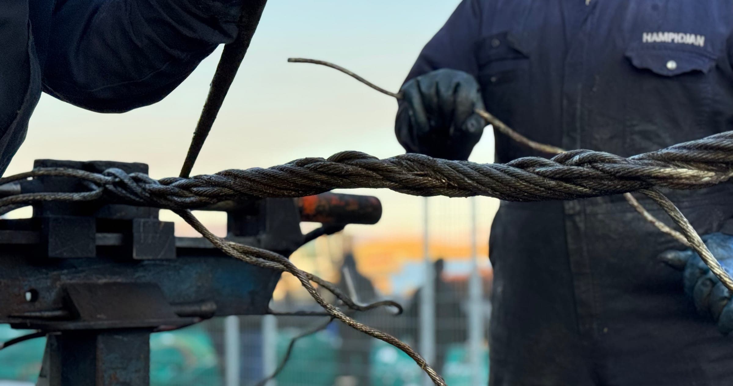 Two riggers hand-splicing a wire rope on the bench, a strand tucked clear