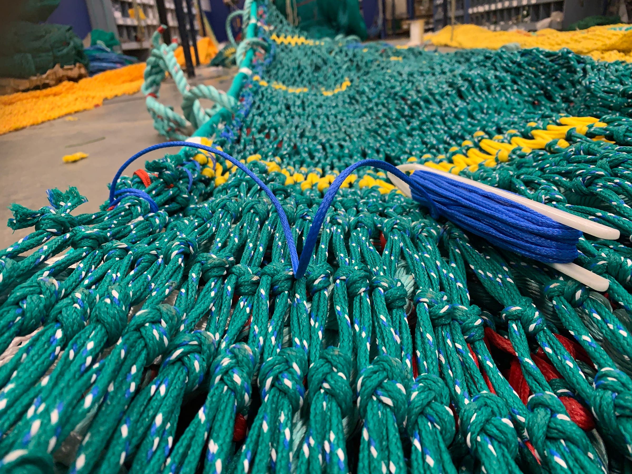 A loaded netting needle and twine laid across green trawl mesh part-way through a repair